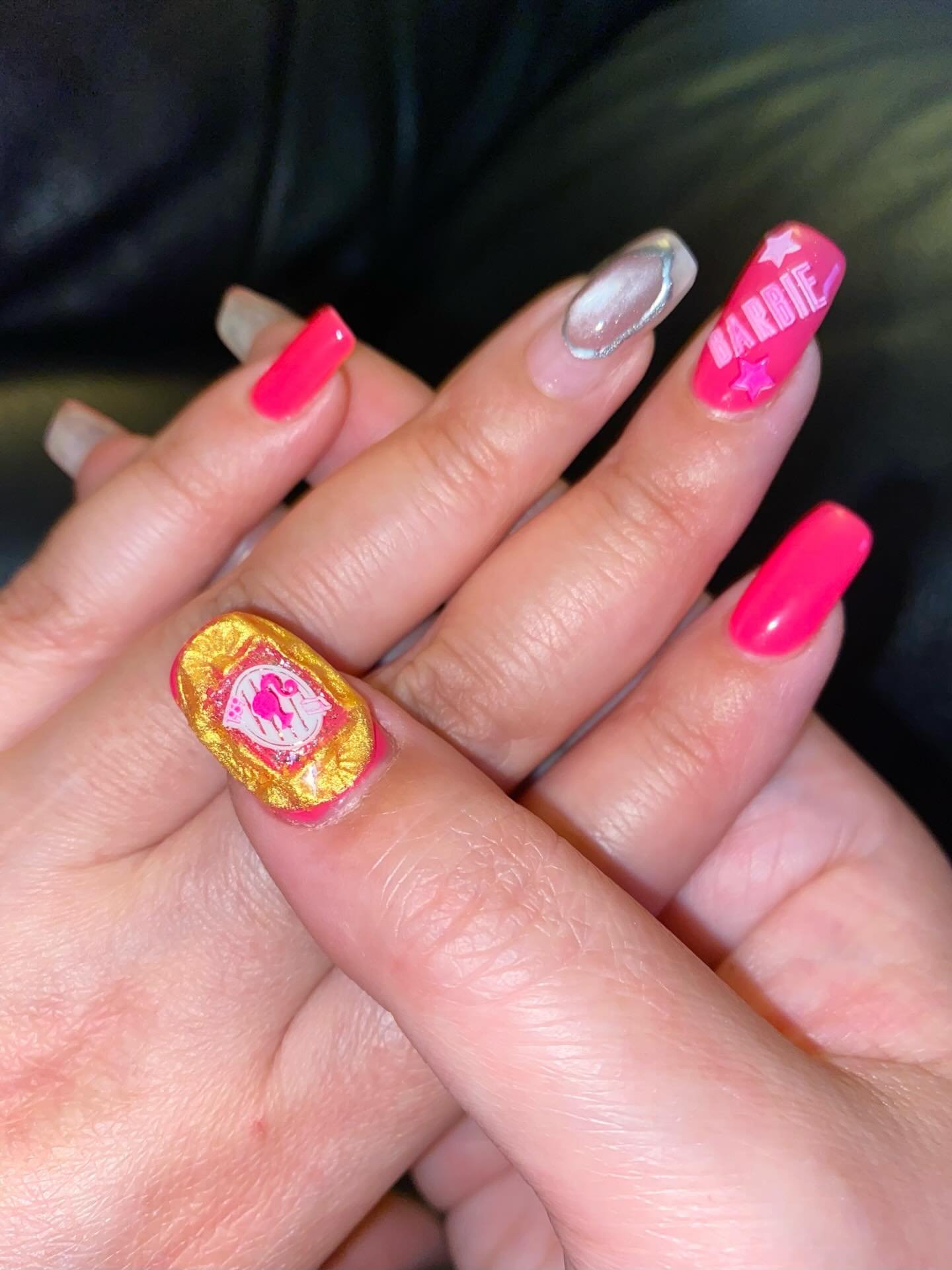 A close-up of a hand displaying trendy coffin-shaped barbie nails with hot pink polish, gold foil accents, and silver details.