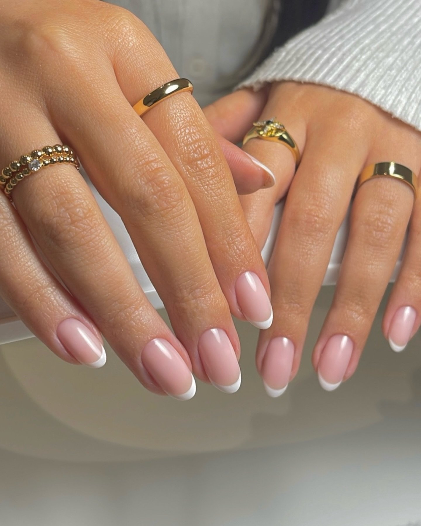 Hands displaying short almond nails ideas with a classic French manicure in soft pink and white, complemented by gold rings for a polished, chic look.