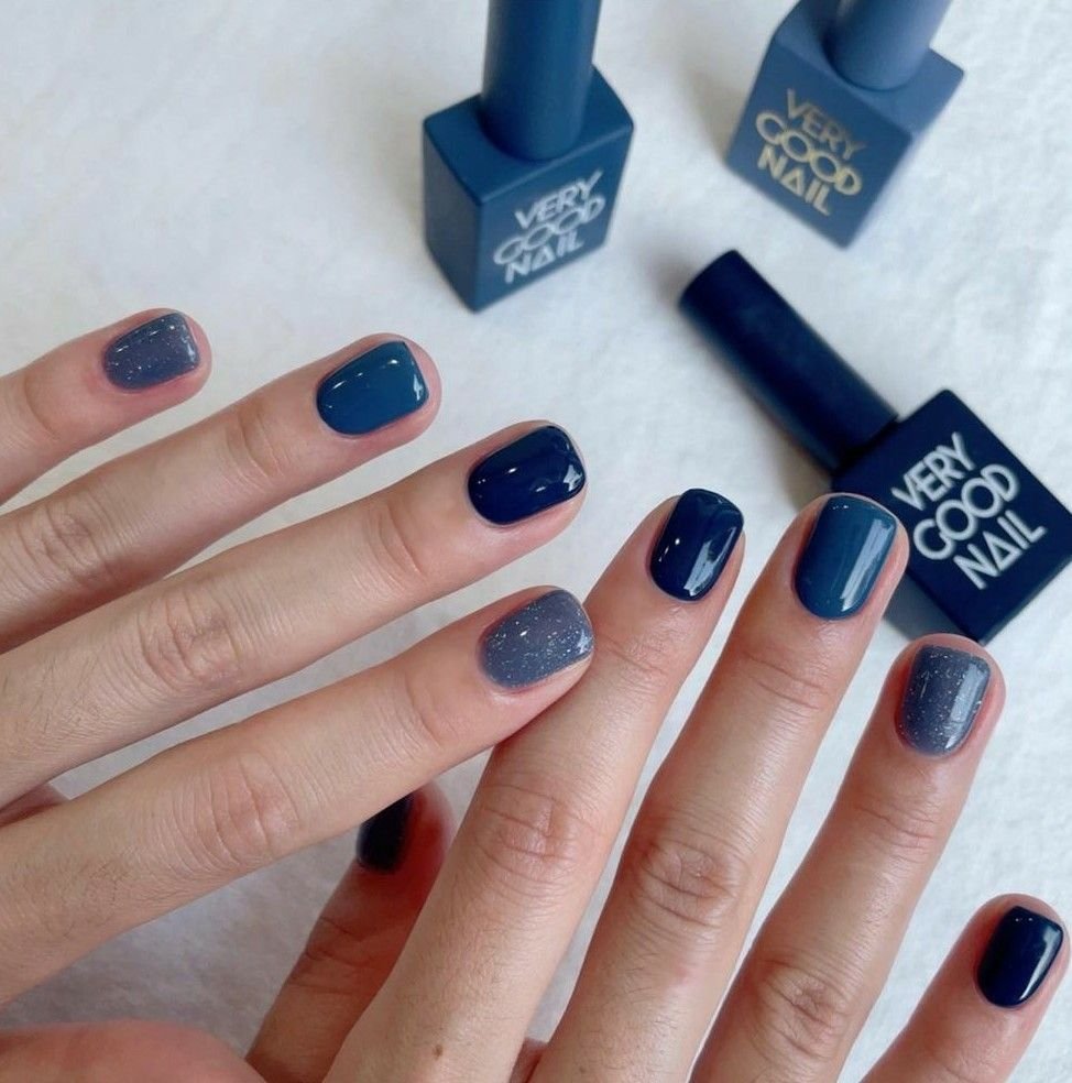 Hand displaying blue short nails ideasle with a mix of deep navy, muted blue, and glittery shades, paired with 'VERY GOOD NAIL' polish bottles on a light surface for a polished, minimalist vibe