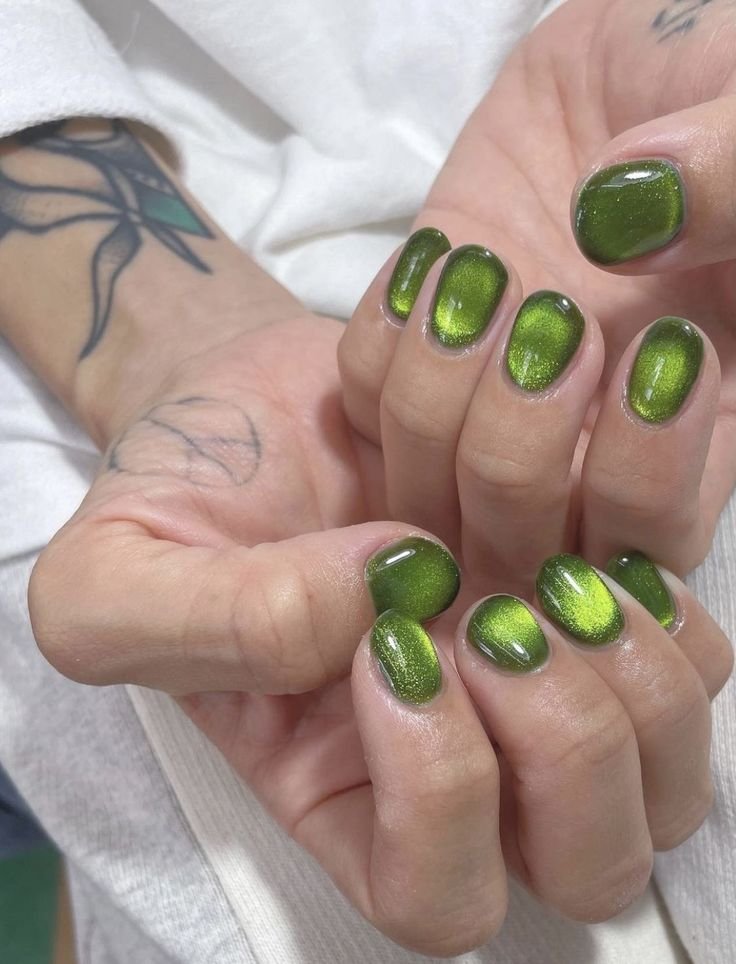 Close-up of short green nails ideas showcasing glossy, magnetic green nail art with a shimmering cat-eye effect, hands positioned on white fabric with a floral wrist tattoo in the background.
