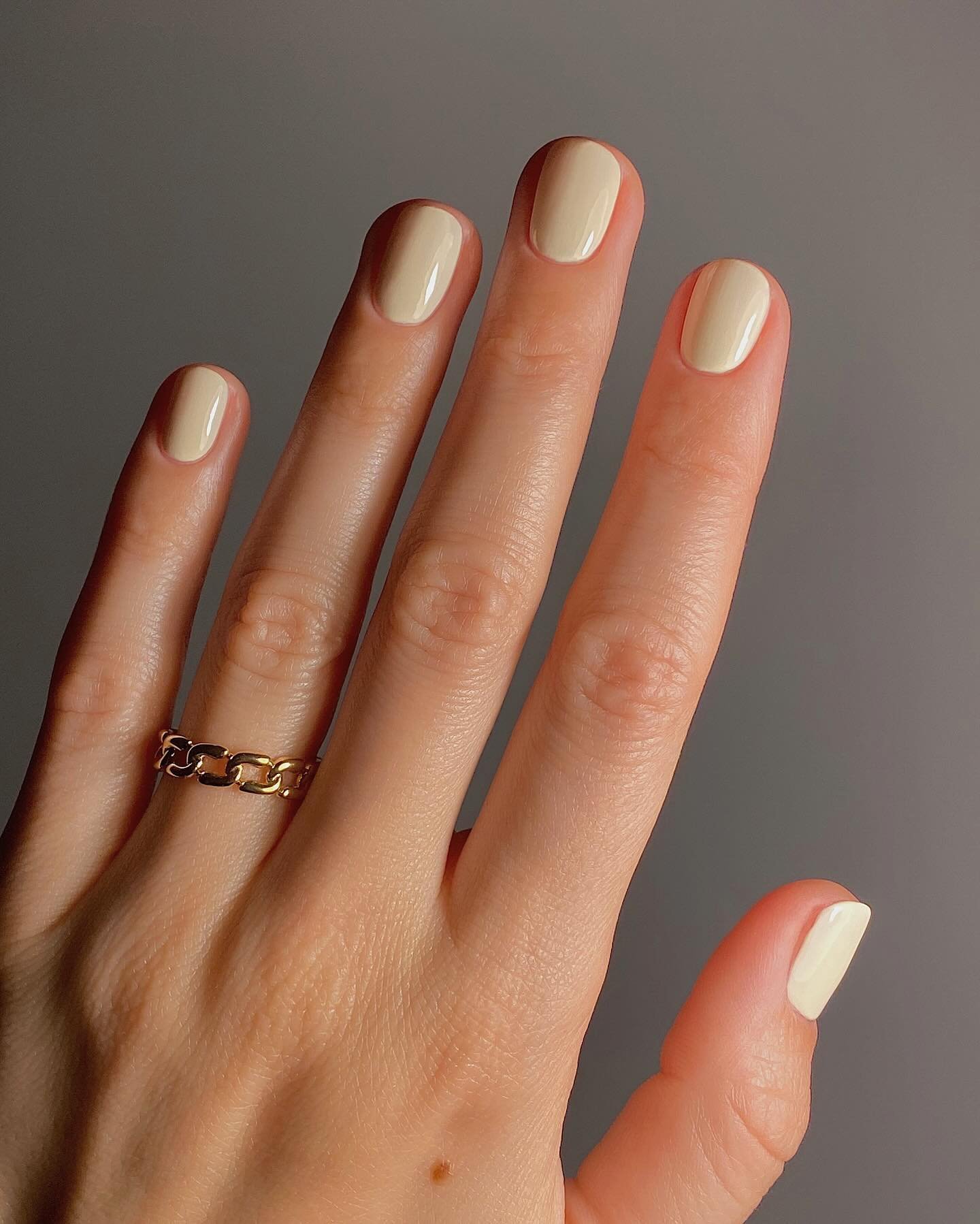 Close-up of a hand showcasing short nails ideas simple with a soft beige manicure, gold chain ring, and neutral gray backdrop for a clean, minimalist aesthetic