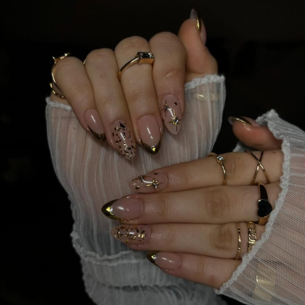 Get inspired by artistic short almond nails ideas incorporating a nude base with elegant gold French tips, adorned with celestial motifs like tiny black stars, birds, and shimmering gold foil for a whimsical and luxurious starry night effect.