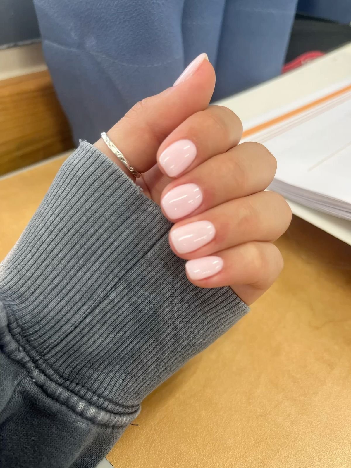 Hand featuring pink short nails ideas with glossy light pink nail polish, a delicate silver ring, and a gray ribbed sleeve, set against a cozy indoor backdrop.