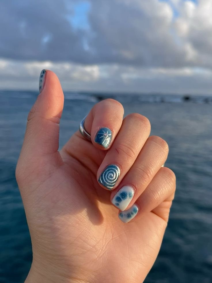 Close-up of a hand showcasing creative short nail ideas for summer with blue oceanic designs including spirals and stars against a scenic sea backdrop.