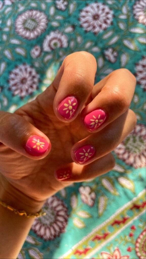 Hand with pink short nail ideas for summer decorated with yellow daisy flowers on a turquoise floral patterned background.