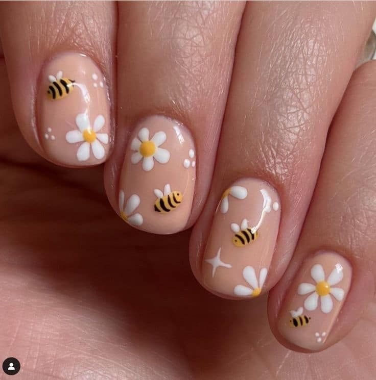 Short spring nails ideas featuring nude polish with white daisy flowers and cute yellow and black bee nail art on rounded short nails.