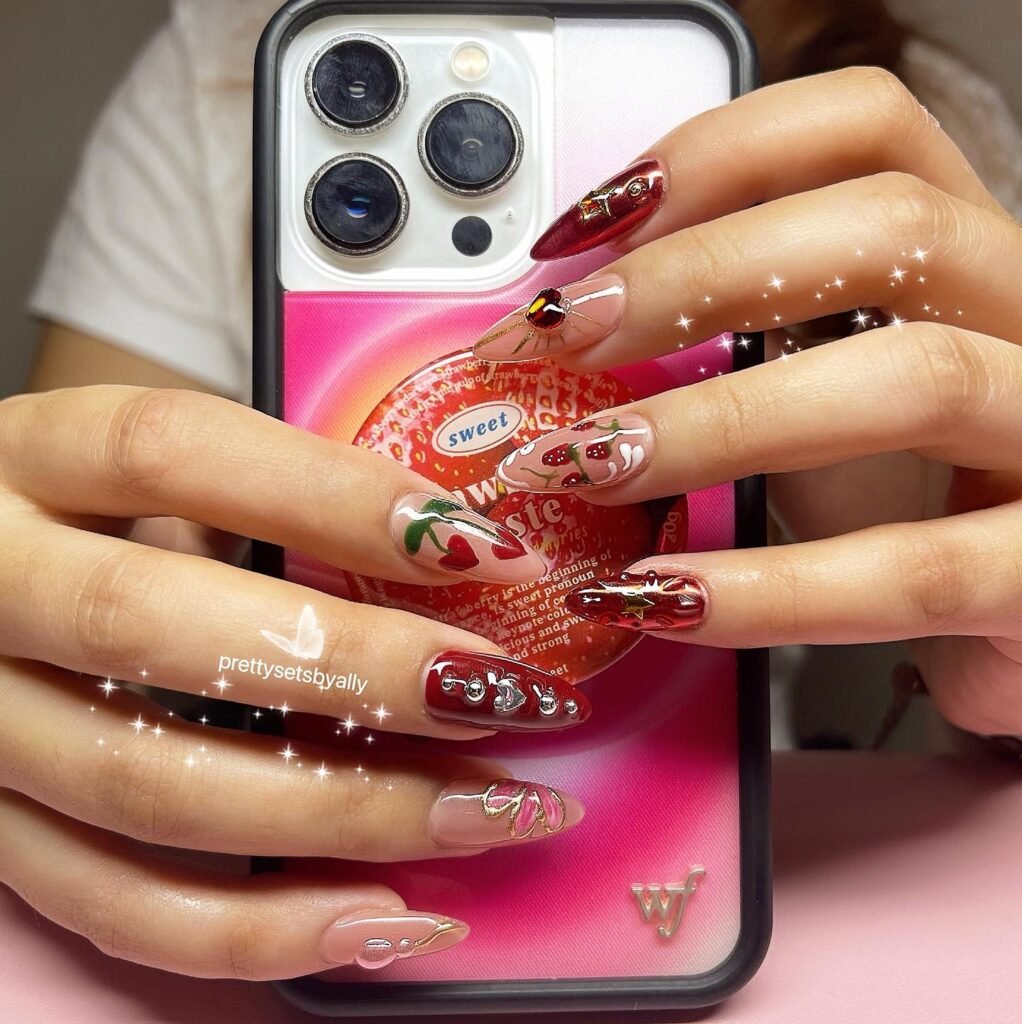 Creative hawaii nails ideas featuring cherry red tones with fruit motifs, jewels, and intricate details on long pointed nails holding a phone.