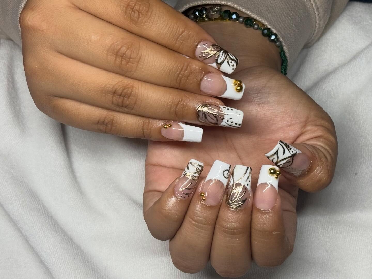 A close-up of long square nails featuring chunky white tips, gold foil leaf accents, and tiny gold bead charms, showcasing a modern take on 2000s French tip nails with Y2K-inspired details.