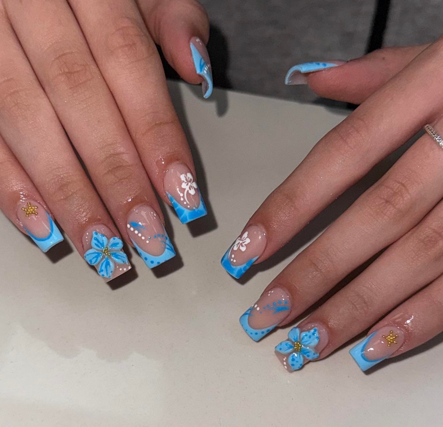 A close-up of a manicure with long, square nails featuring light blue French tips. The design showcases 3d french tip nails embellished with raised blue flowers, white dots, and gold star charms.