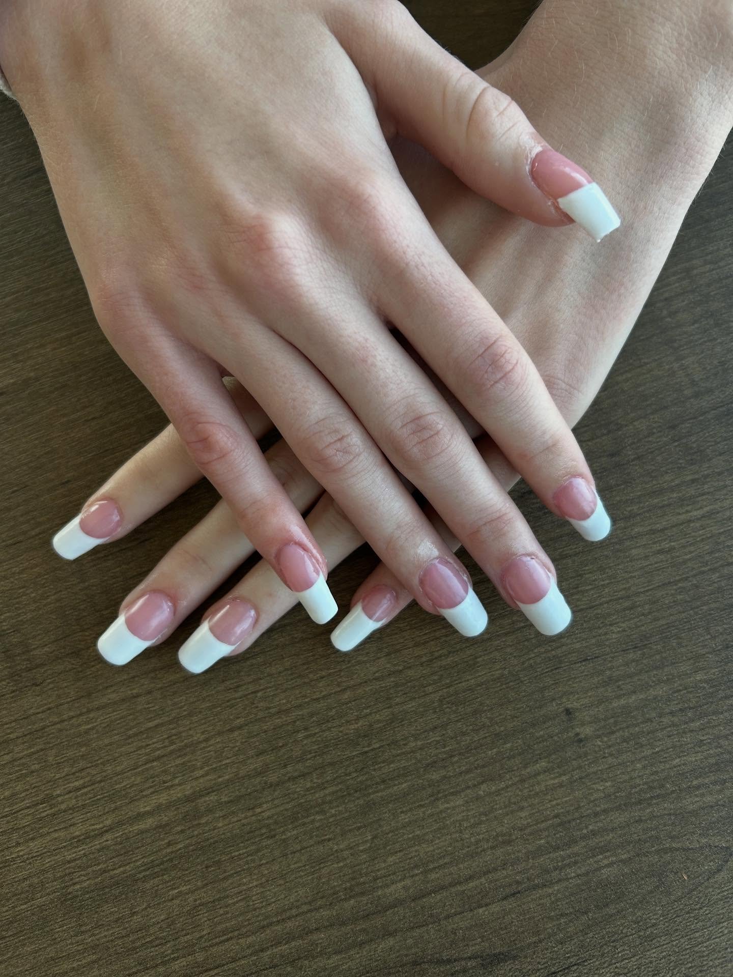 A pair of hands resting on a wooden surface displaying short, square nails with classic thick white tips and a nude pink base, perfectly capturing the timeless aesthetic of 90s French Tip Nails.