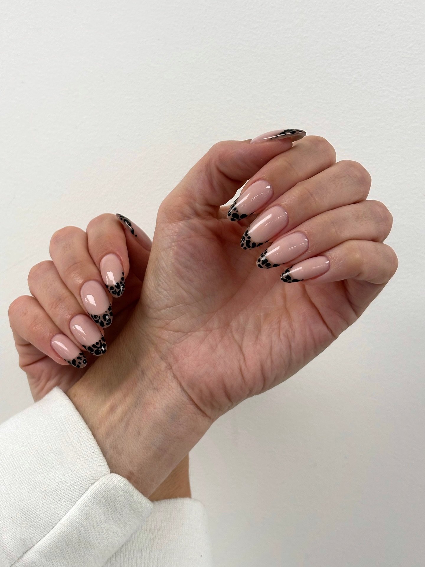 A pair of hands against a white background featuring long almond nails with a sheer nude base and black spotted tips, presenting a chic and modern style of Cheetah Print French Tip Nails.