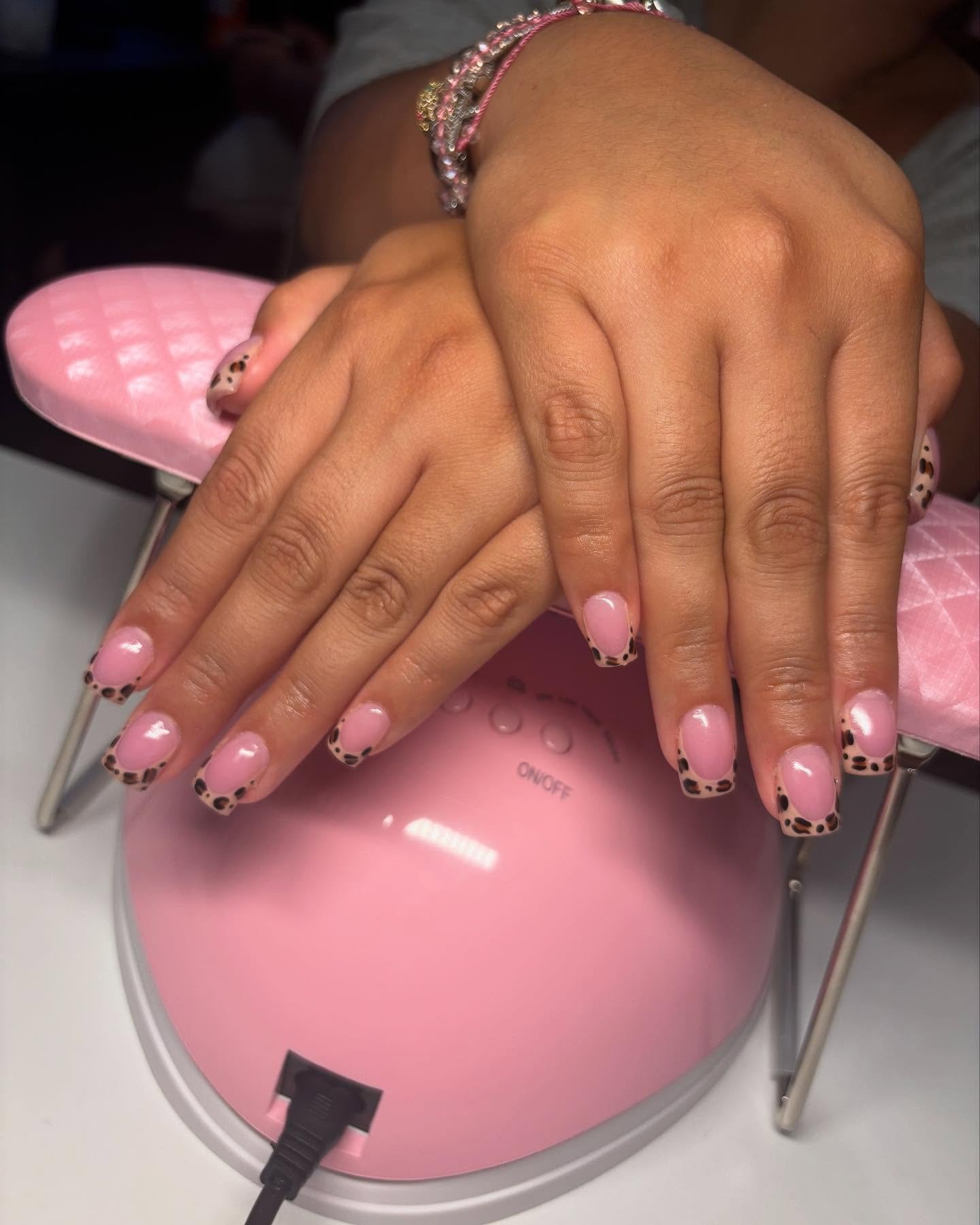 Hands resting on a pink nail lamp displaying square-shaped nails with a glossy pink base and animal print tips, showcasing playful Cheetah Print French Tip Nails.