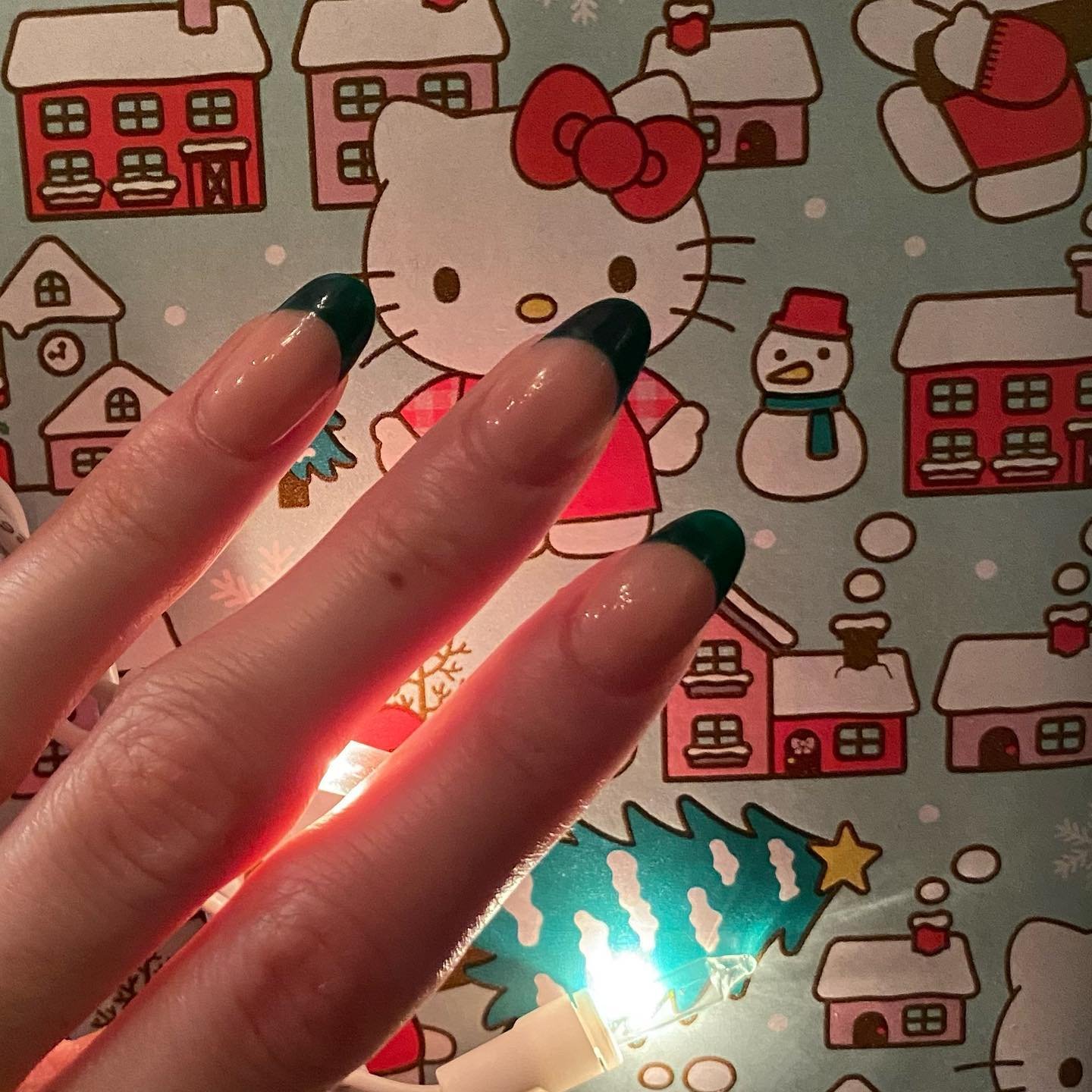 Three fingers displaying medium-length green french tip nails in a solid deep emerald shade, set against a festive Hello Kitty Christmas background with warm fairy lights.