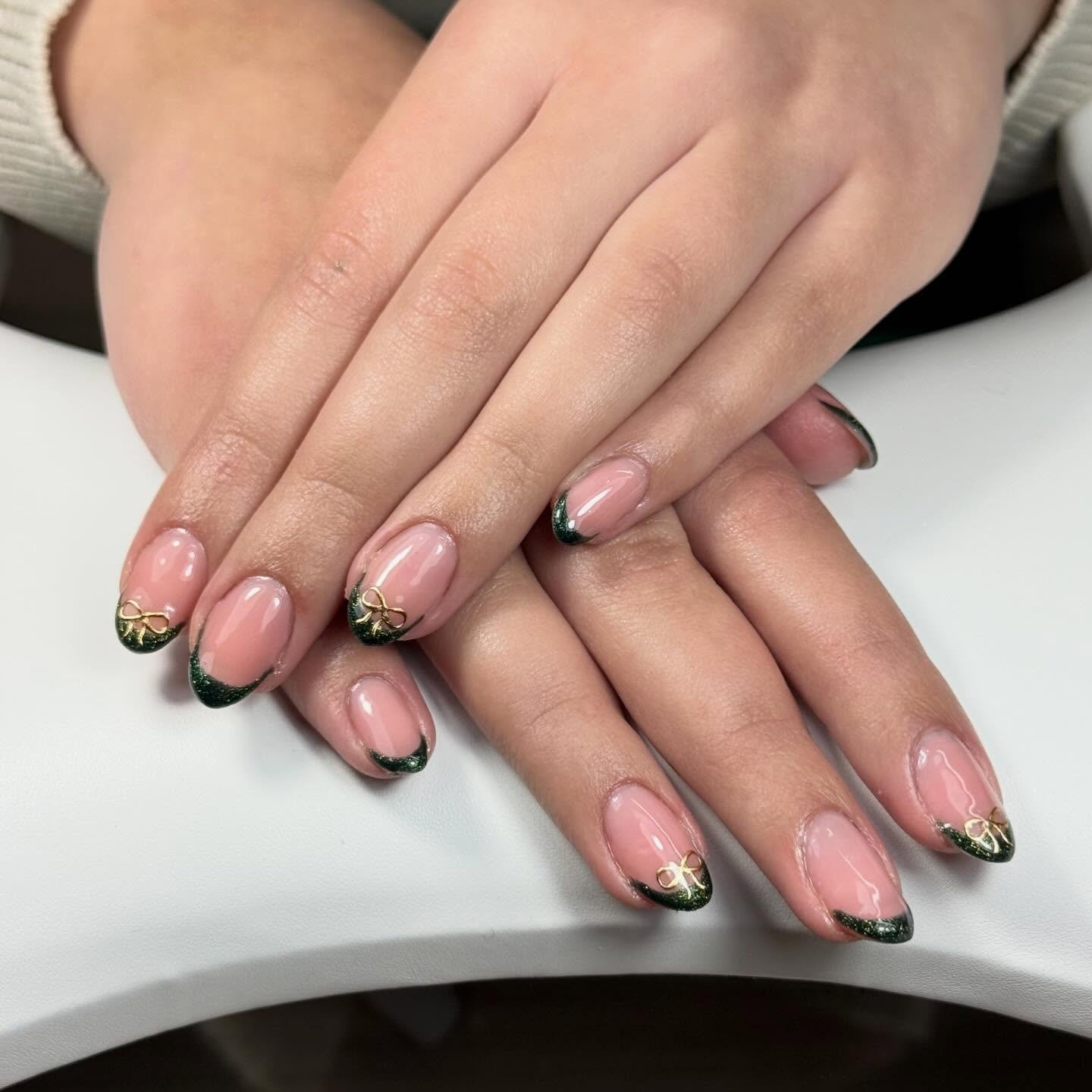 Hands with almond-shaped nails featuring a sheer pink base and dark green glitter, showcasing elegant green french tip nails accented with delicate gold bow charms.