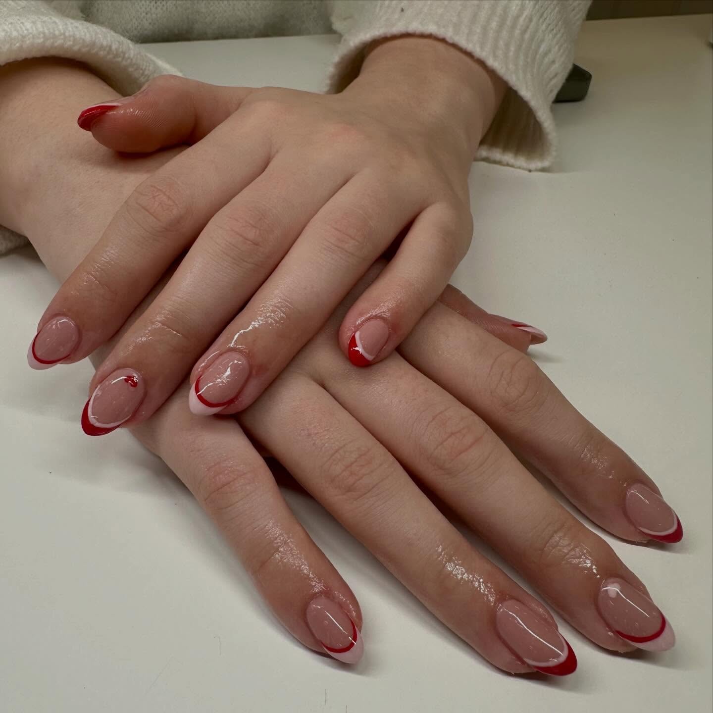 Hands displaying short, rounded nails with a nude base, red tips, and delicate white inner lines, illustrating a chic style of double french nails with subtle heart accents.