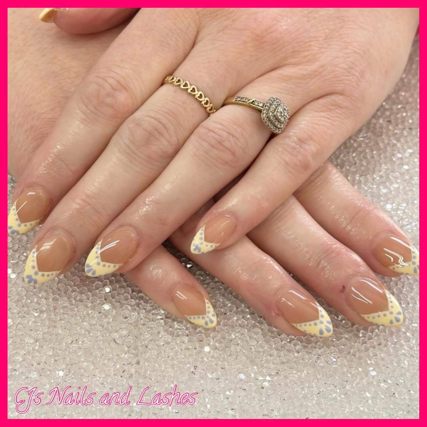 A hand adorned with gold rings displaying long almond nails with a nude base and pale yellow tips decorated with blue dots, showcasing a playful take on the glazed french tip nails trend with a high-shine finish.