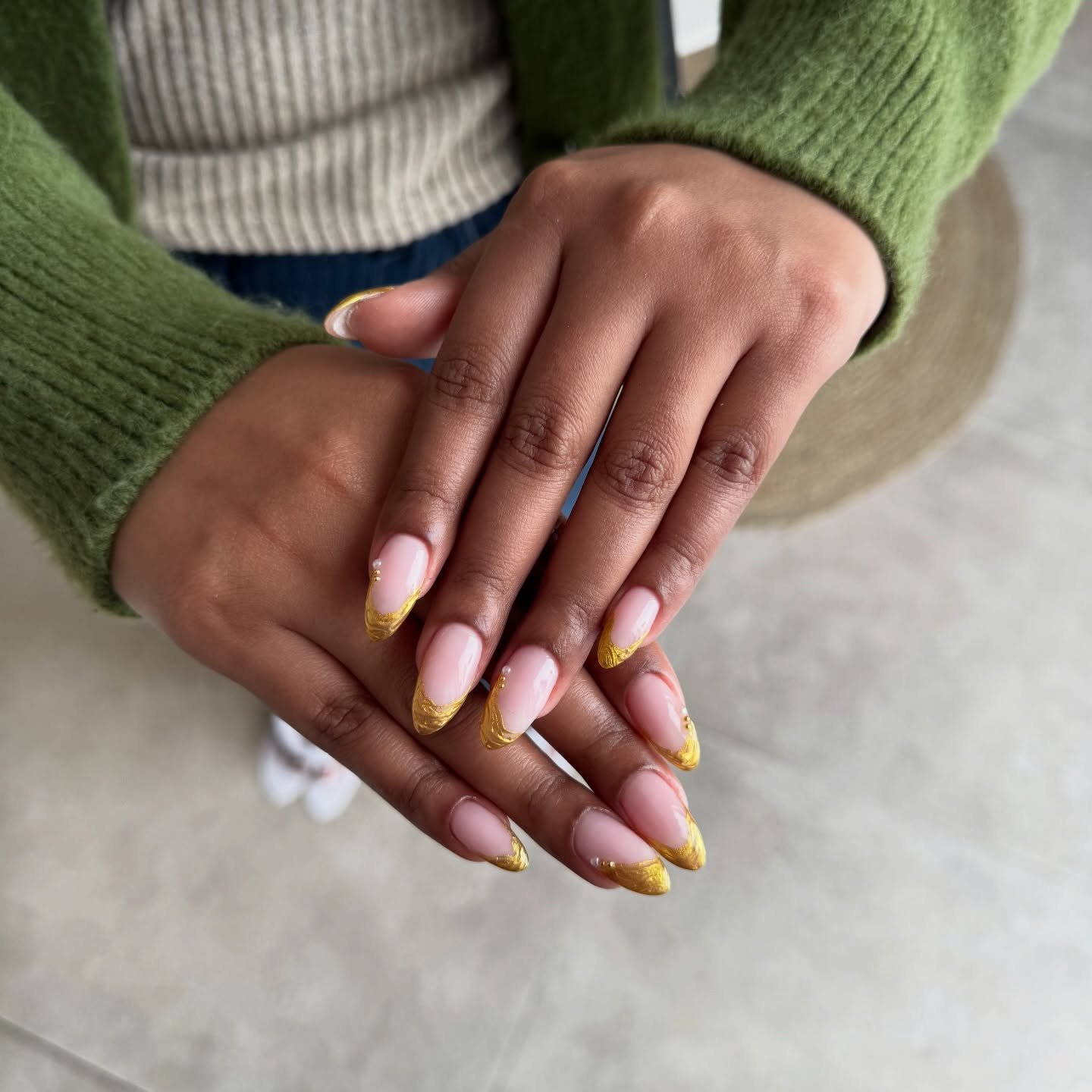 Hands wearing a green sweater displaying long almond nails with a milky pink base and textured gold tips, presenting a glamorous style of Gold French Nails with pearl accents.