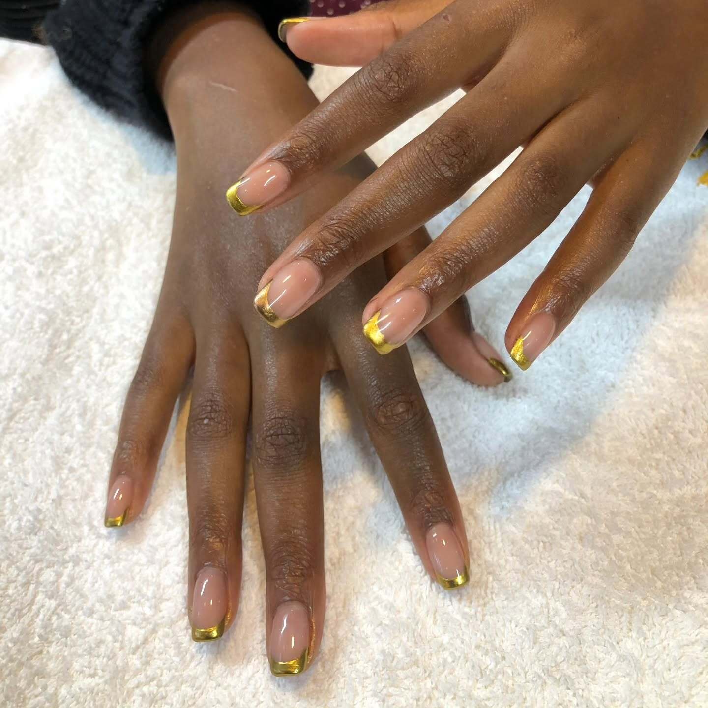 A close-up of hands with square-shaped nails featuring a sheer nude base and bold metallic tips, showcasing classic Gold French Nails against a white towel.