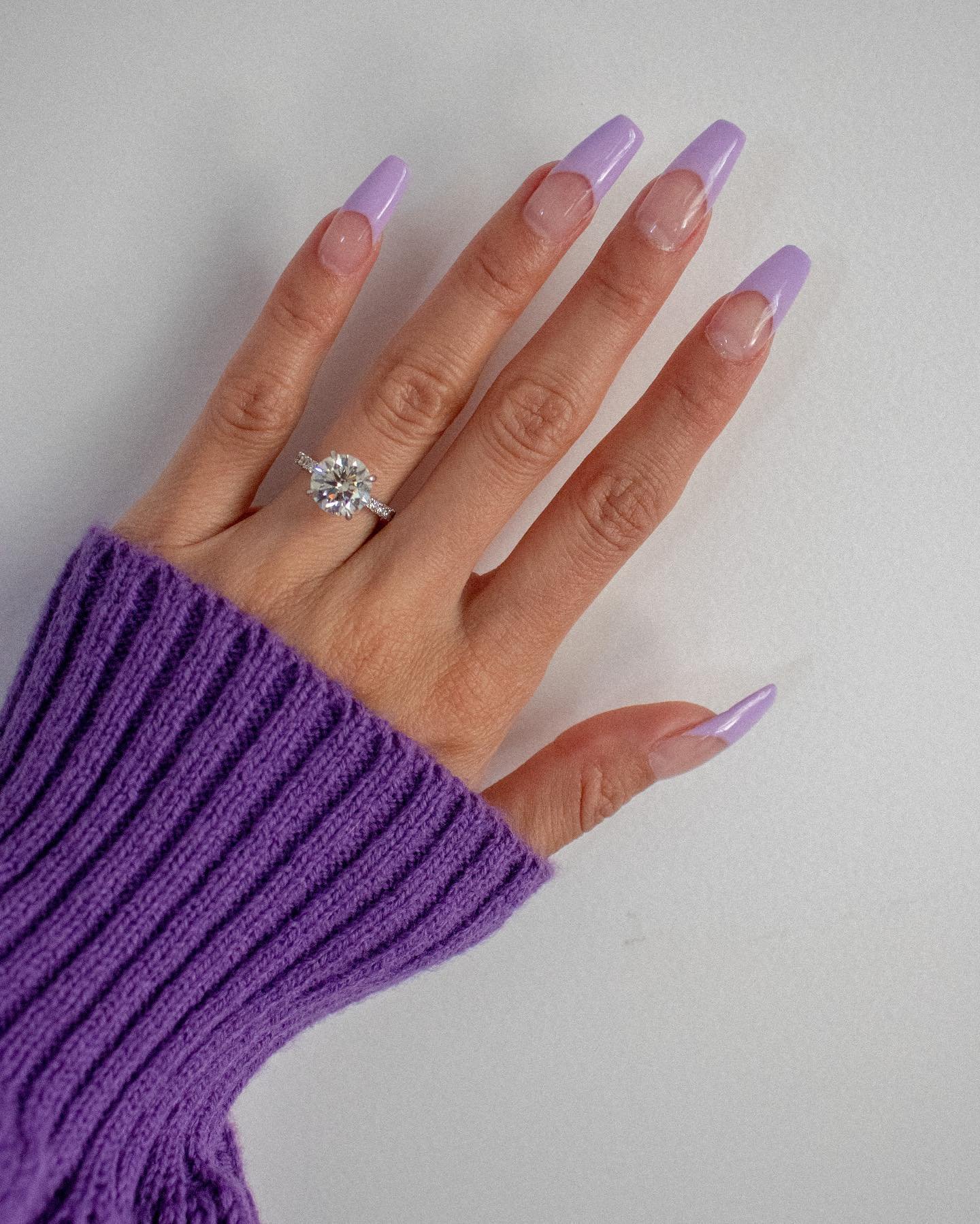 A hand wearing a purple sweater and a diamond ring displays long coffin nails with a sheer base and soft purple tips, exemplifying elegant lilac french tip nails.