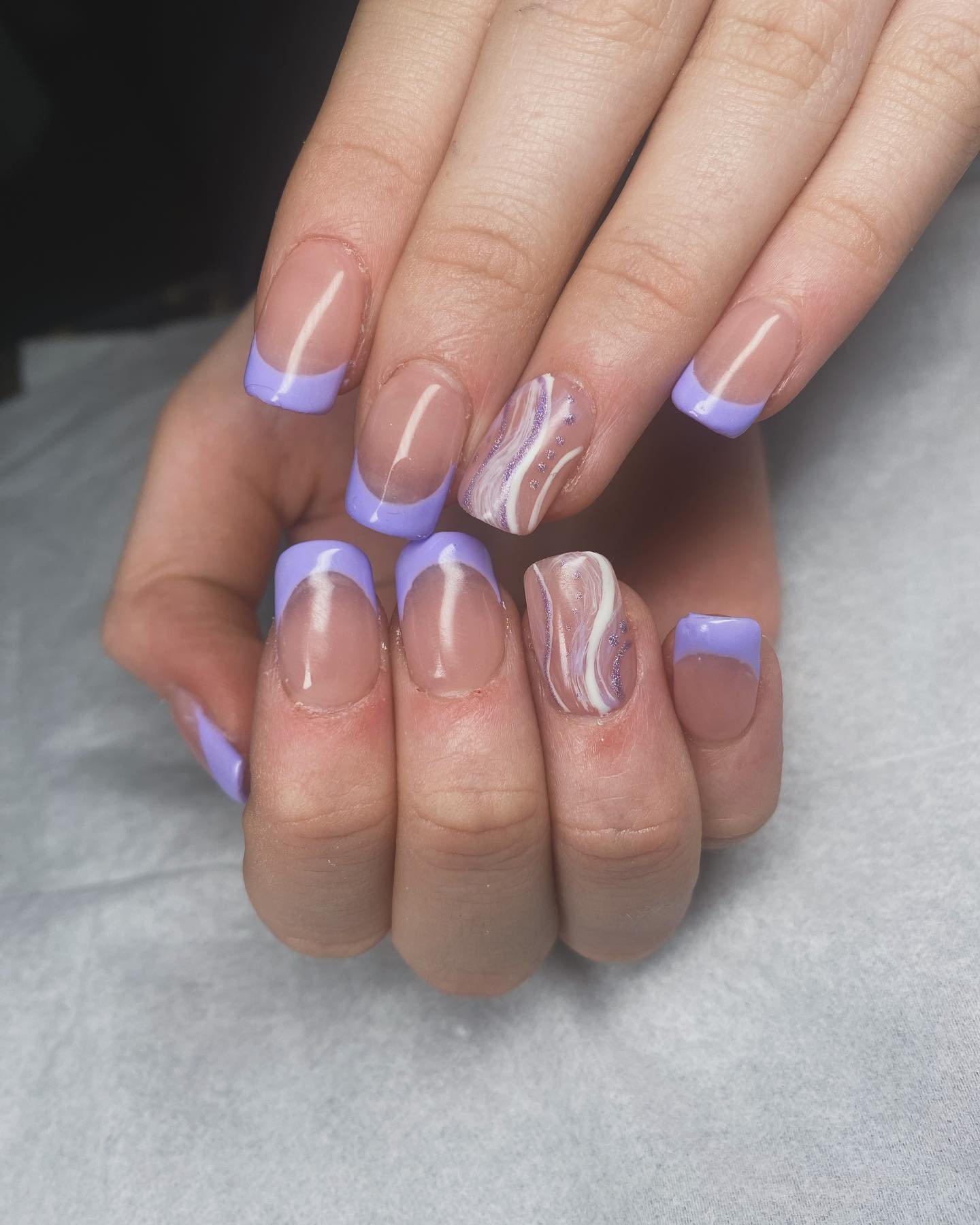 A close-up of short square nails with a nude base and light purple tips, including accent nails with white marble swirls, showcasing artistic lilac french tip nails.