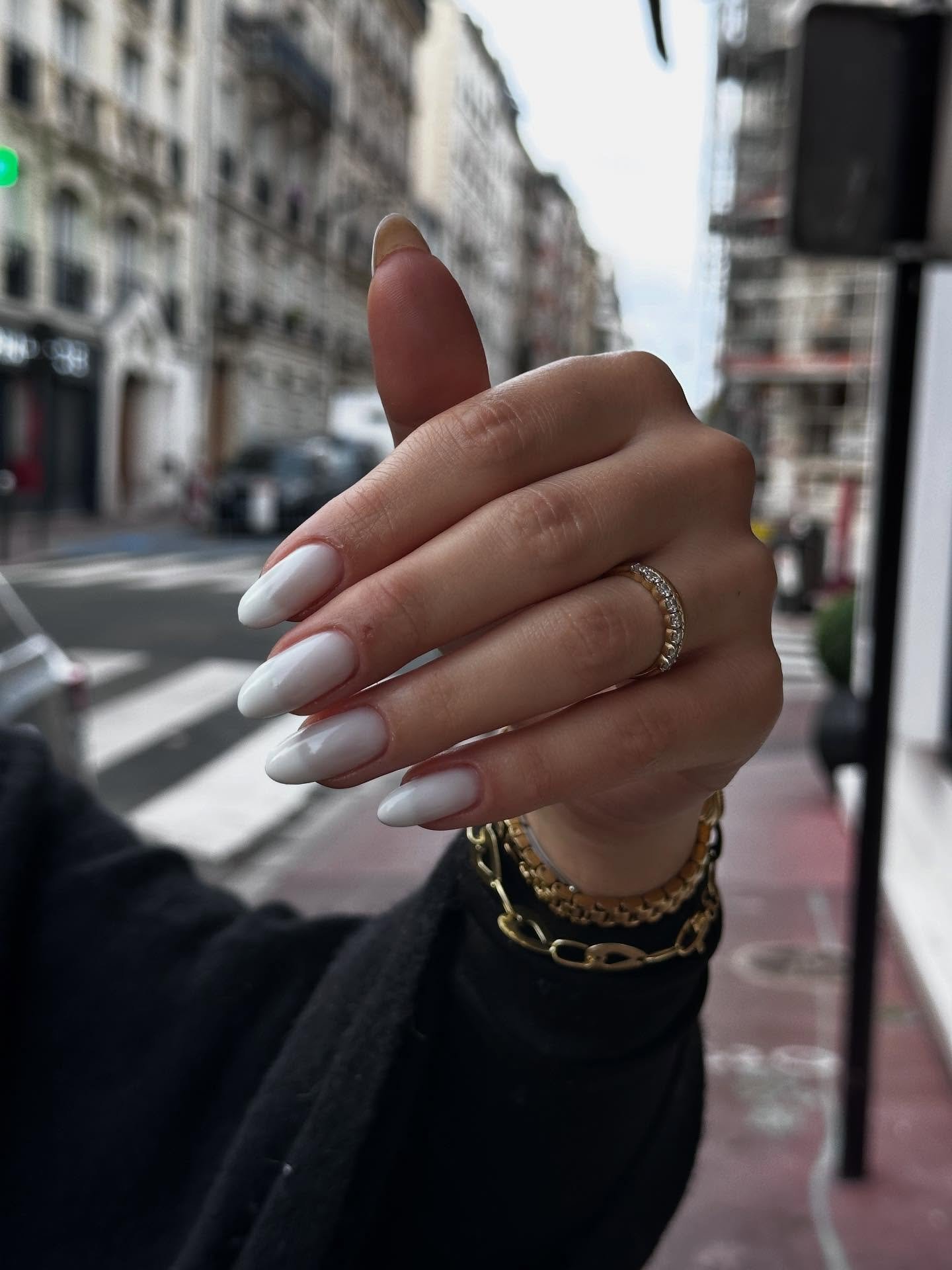 A hand held up against a blurred city street background displaying long, almond-shaped milky white nails and gold jewelry, showcasing a modern variation of natural french tip nails.