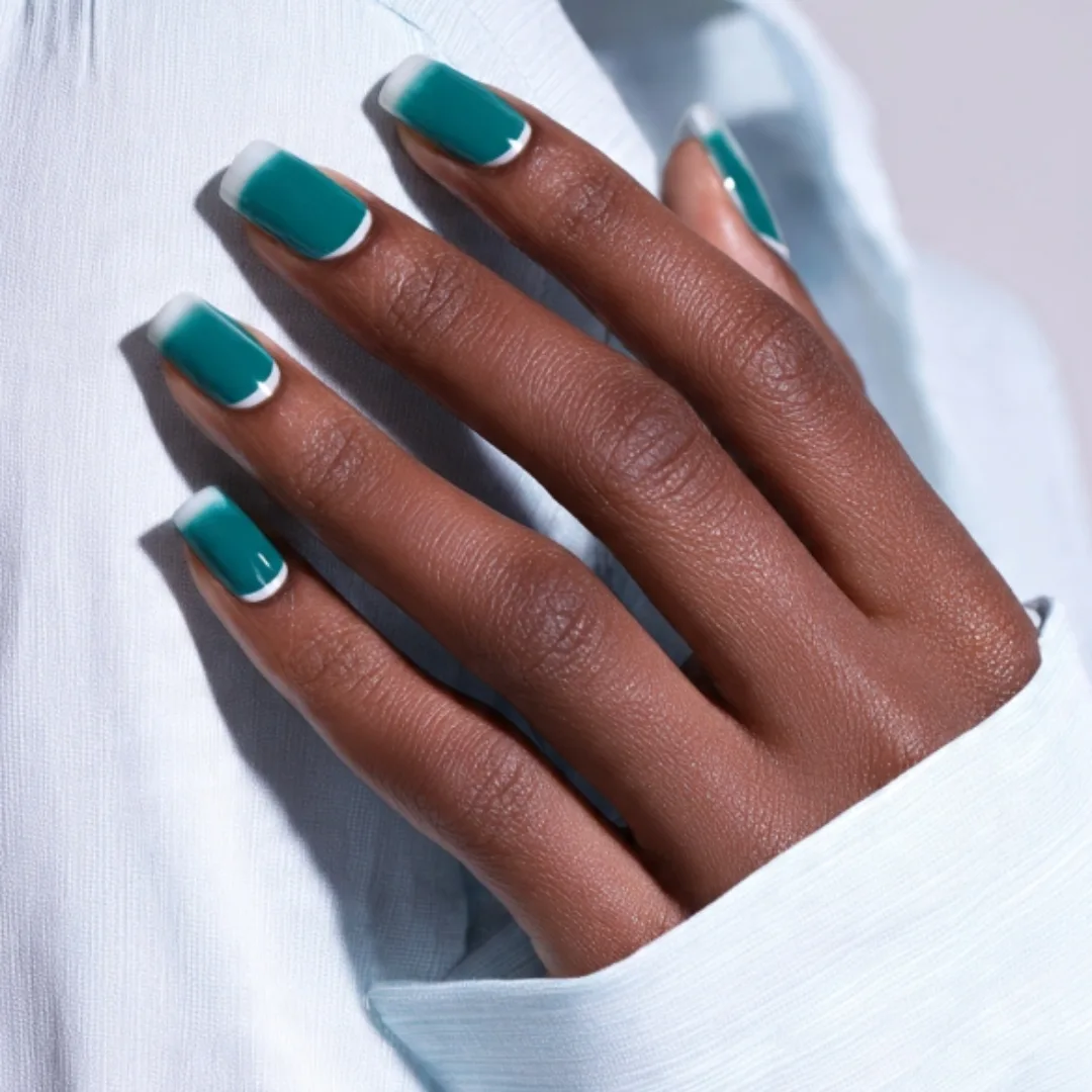 A hand resting on a white shirt displaying square-shaped nails with a deep teal base and white tips, showcasing a classic style of teal french tip nails.
