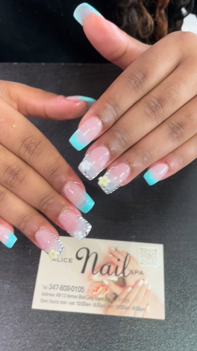 A close-up of long square nails featuring bright teal french tip nails, alternating with white tips decorated with rhinestones and 3D flowers, posed over a nail salon business card.