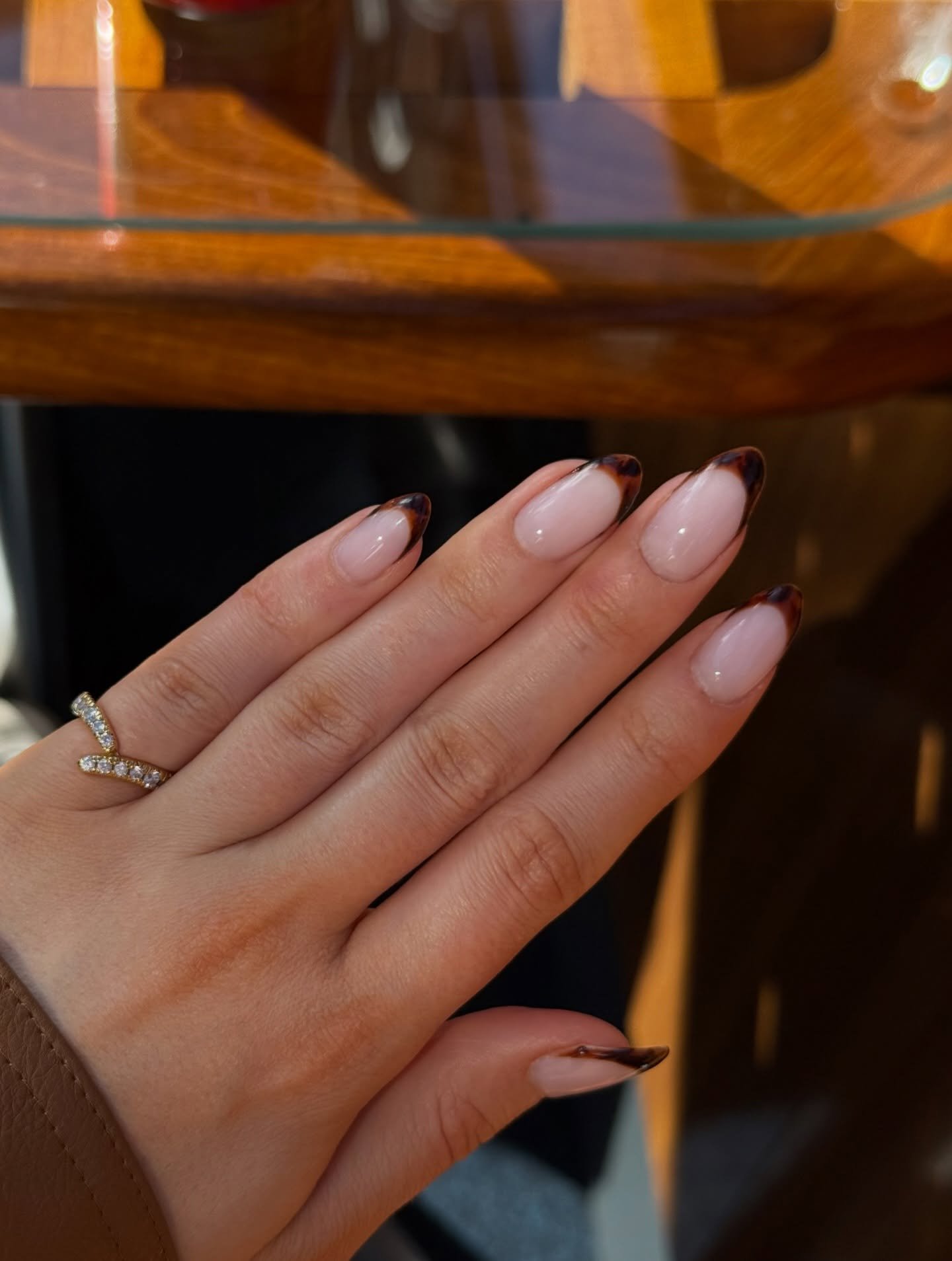 A hand resting near a wooden table displaying long almond-shaped nails with a soft pink base and warm amber tips, presenting a polished take on tortoise shell french tip nails accented by a delicate diamond ring.