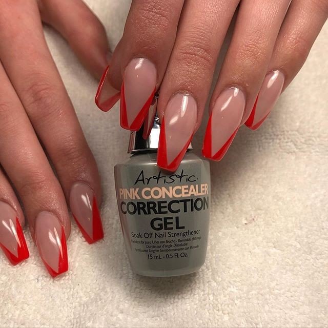 A close-up of long stiletto nails with a glossy nude base and bold red v shaped french tip nails, styled around a bottle of Artistic Pink Concealer gel.