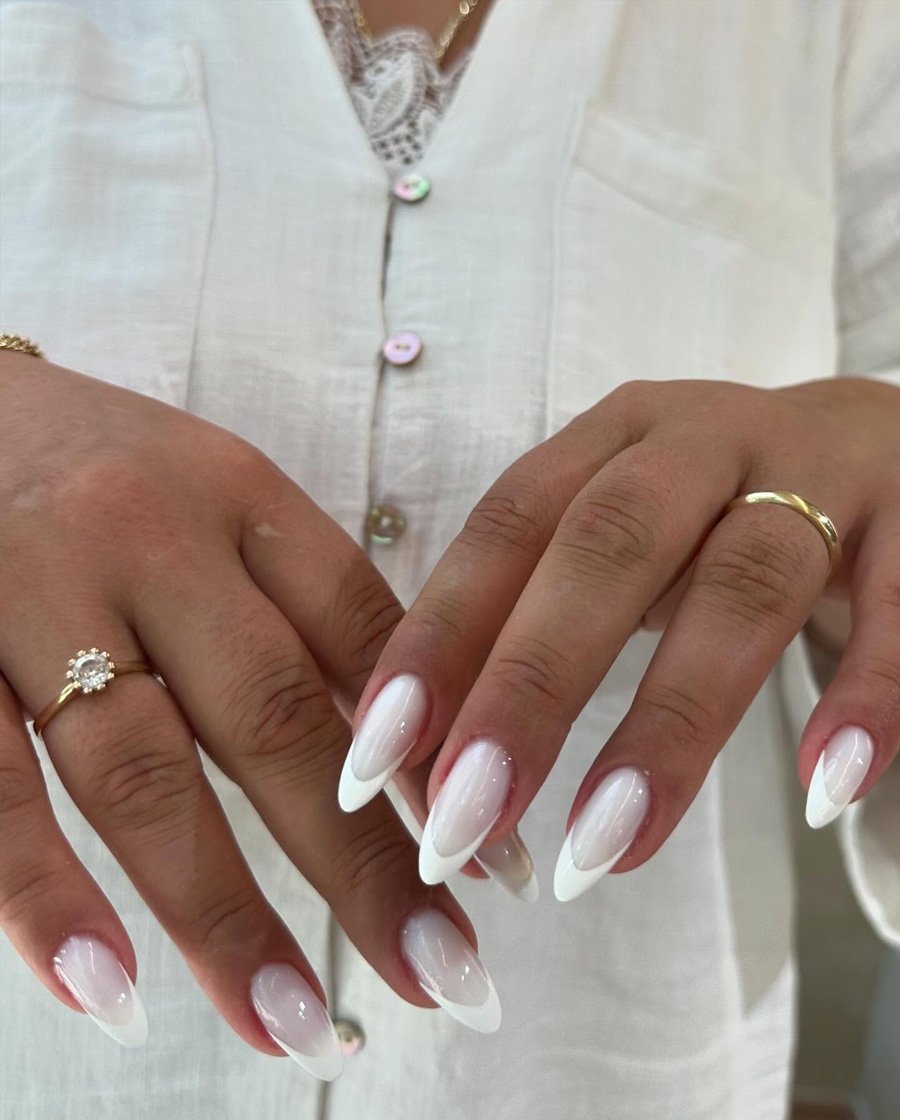 A close-up of long almond-shaped nails featuring a soft pink base with crisp white tips, showcasing elegant white french nails paired with gold rings against a cream-colored outfit.