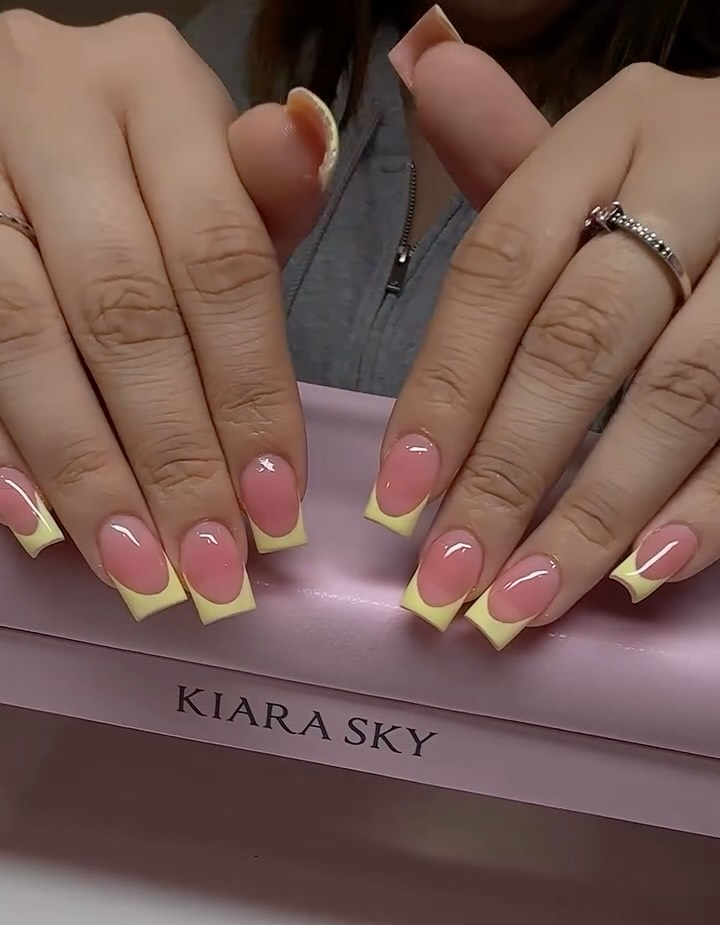 A close-up of square-shaped nails with a glossy pink base and bright yellow tips, showcasing cheerful yellow french tip nails displayed over a pink Kiara Sky box.