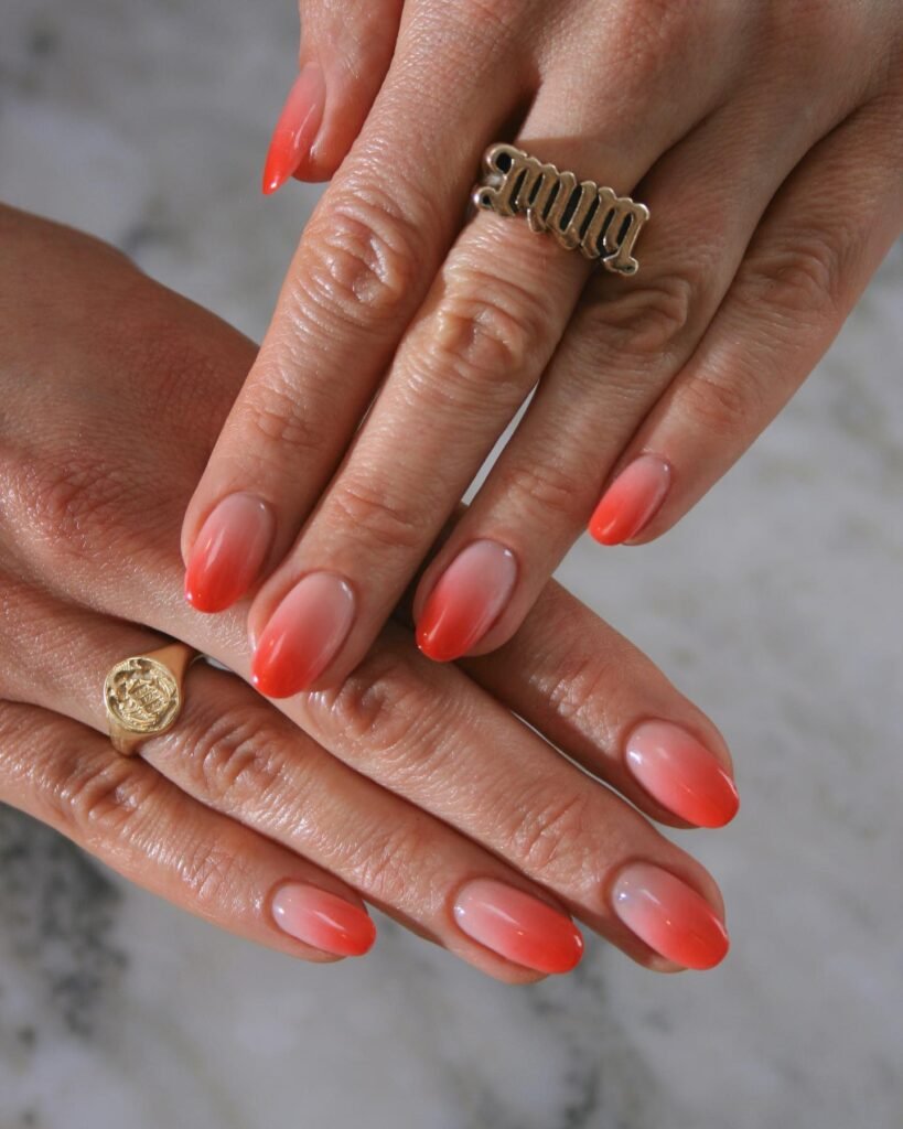 A close-up of hands displaying almond-shaped nails with a coral and nude gradient design, perfect for stylish August Nails Ideas, accessorized with gold rings.
